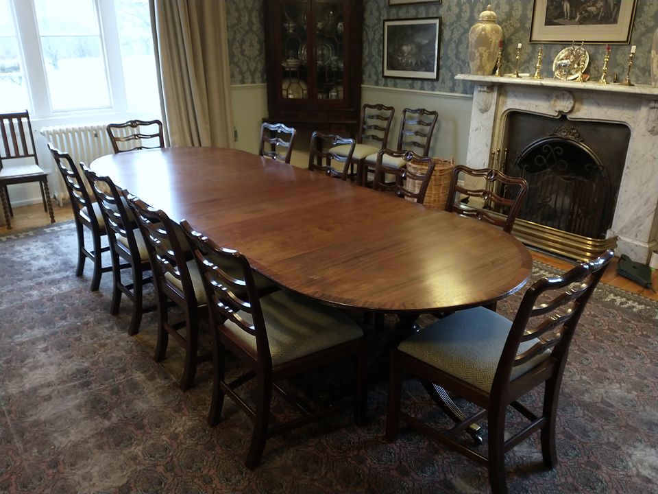 Restored D-end extending dining table with ladder-back chairs in a damask-papered dining room beside a marble fireplace
